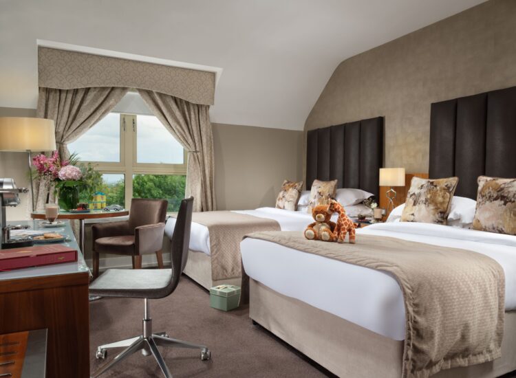 Interior view of a family hotel room with two large beds. Each bed is styled with high vertical headboards, beige runners, and patterned accent pillows. On the closest bed, a stuffed teddy bear and a giraffe toy are neatly arranged. A work desk with an office chair occupies the foreground, while a seating area with a brown armchair and a window showing green trees is visible in the background.
