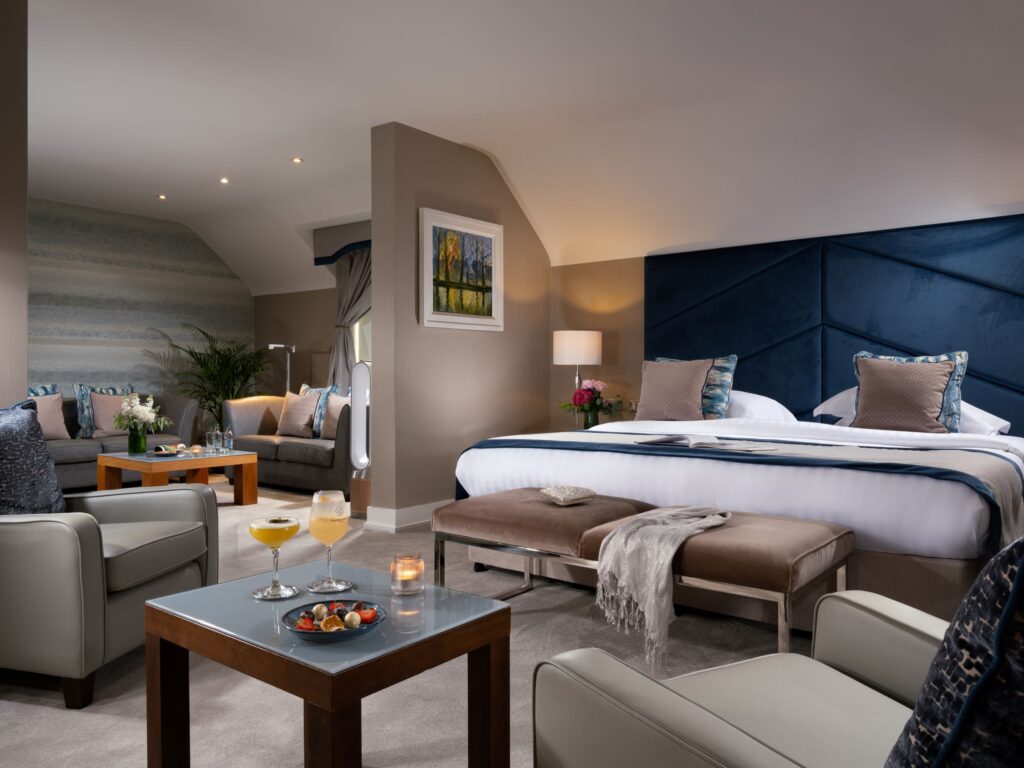 A modern luxury hotel suite with a king-size bed, a deep blue headboard, and a separate living area.