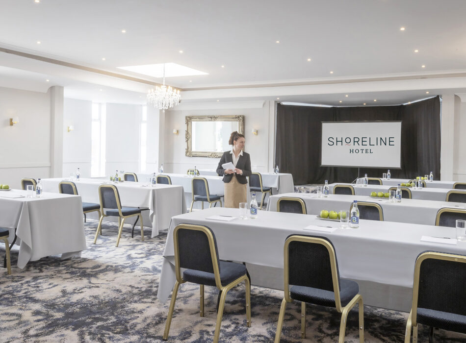 A well-lit hotel conference room set up in a classroom format with long, white-clothed tables, dark blue cushioned chairs with gold frames, and a projection screen displaying "SHORELINE HOTEL" at the front where a woman in professional attire stands.