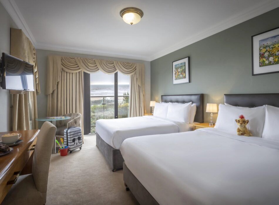 A brightly lit family-friendly hotel room featuring two large beds with white linens, a small plush toy resting against the pillows of the closest bed, a wooden desk with coffee cups, and large glass doors in the background offering a scenic view of the ocean.