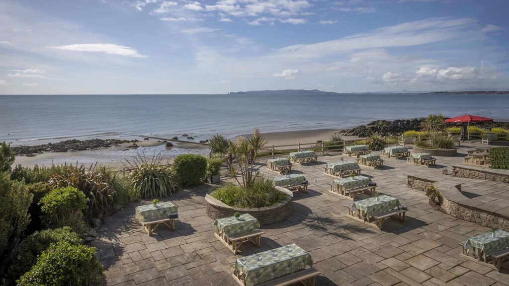 Elevated view of a spacious stone patio featuring several wooden picnic tables covered in patterned tablecloths. The patio is surrounded by lush green shrubs and small palm trees, looking out over a wide sandy beach, calm ocean waters, and a distant coastline under a blue sky with soft white clouds.