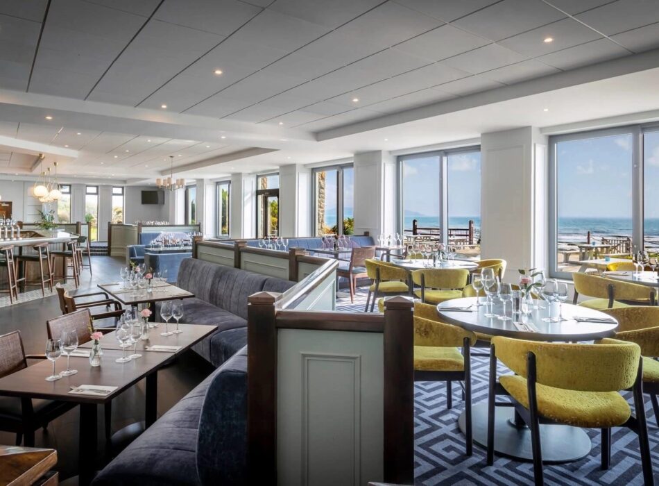 A spacious, modern hotel restaurant and bar featuring a long bar area with stools, dark wood dining tables paired with plush blue banquettes, and round tables with bright yellow chairs arranged next to large floor-to-ceiling windows overlooking a sunny beach and ocean.