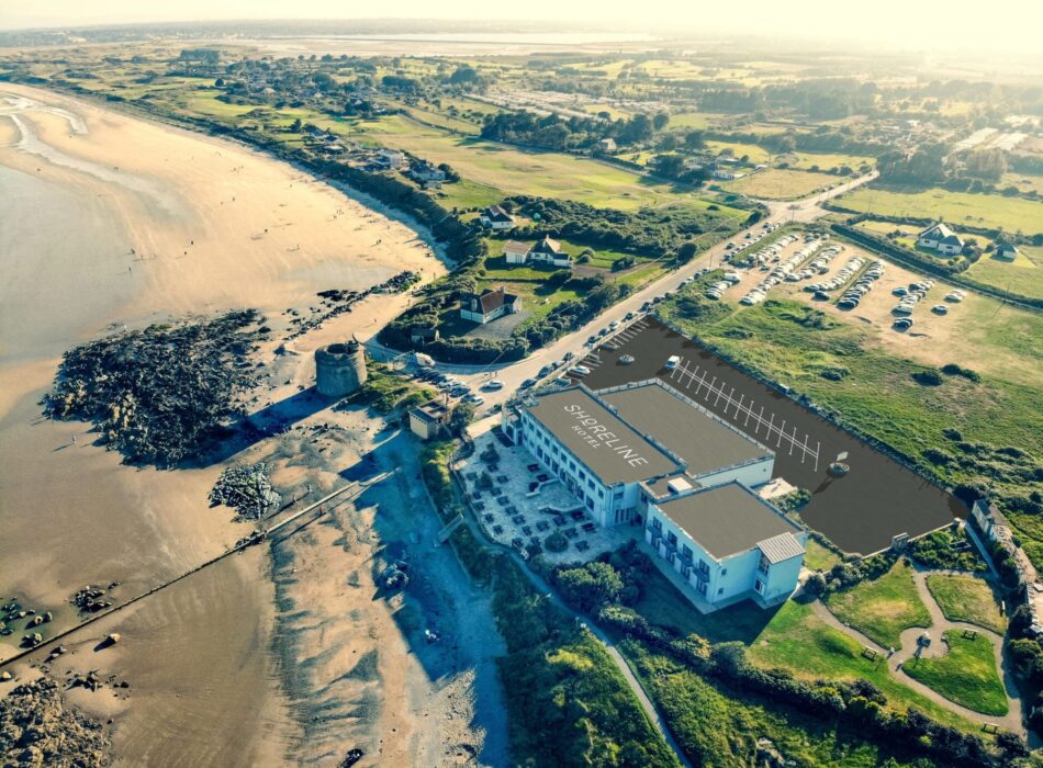 An aerial drone photograph shows the Shoreline Hotel situated directly on a sandy beachfront coastline, featuring a modern multi-story building with its name printed on the roof, an adjacent outdoor patio, an expansive dark asphalt parking lot, and a historic stone Martello tower nearby, all surrounded by green fields and a coastal road under bright daylight.