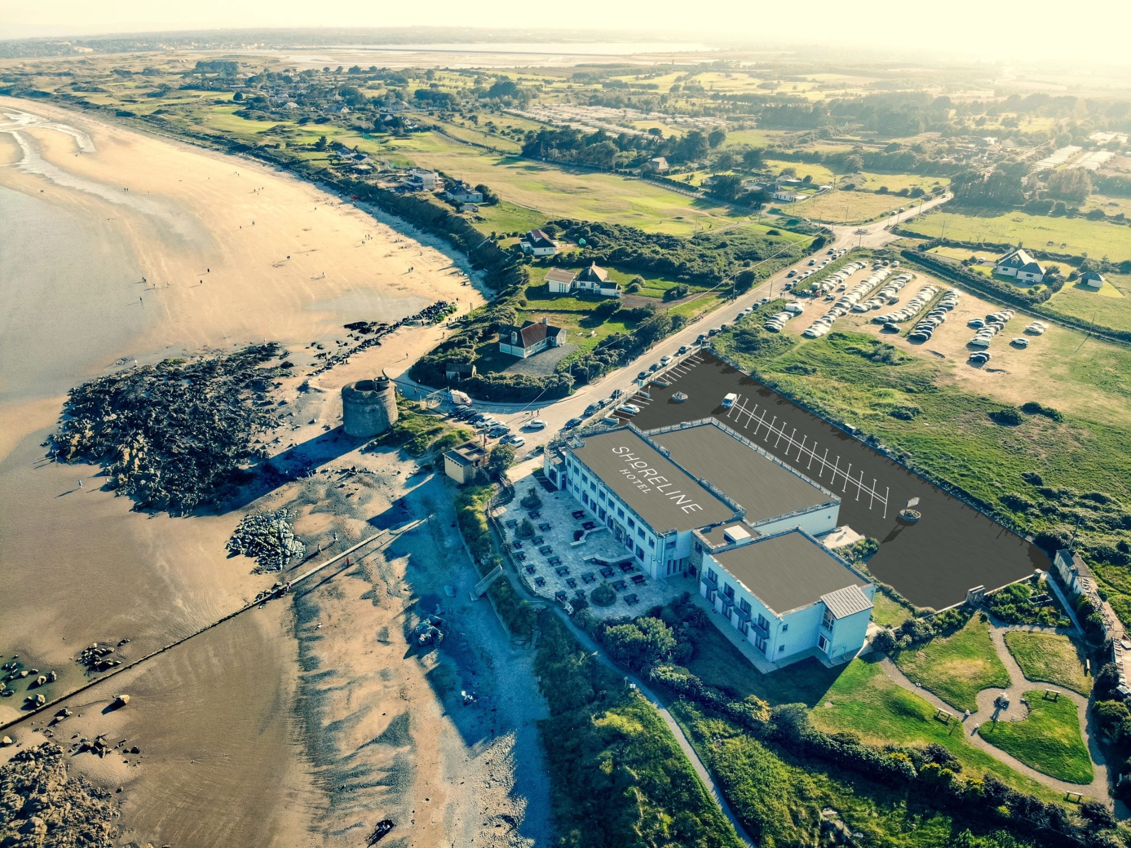 An aerial drone photograph shows the Shoreline Hotel situated directly on a sandy beachfront coastline, featuring a modern multi-story building with its name printed on the roof, an adjacent outdoor patio, an expansive dark asphalt parking lot, and a historic stone Martello tower nearby, all surrounded by green fields and a coastal road under bright daylight.
