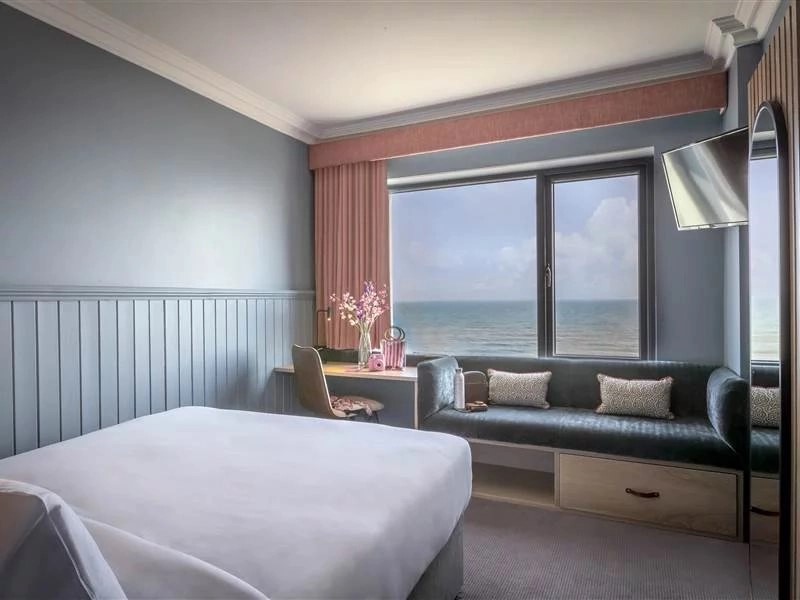 A brightly lit, modern hotel room interior featuring a neatly made bed with crisp white linens in the foreground, light blue walls with matching vertical wood paneling, a dark built-in sofa under a large window that frames a wide view of the ocean, pink curtains, and a small writing desk with a chair in the corner.