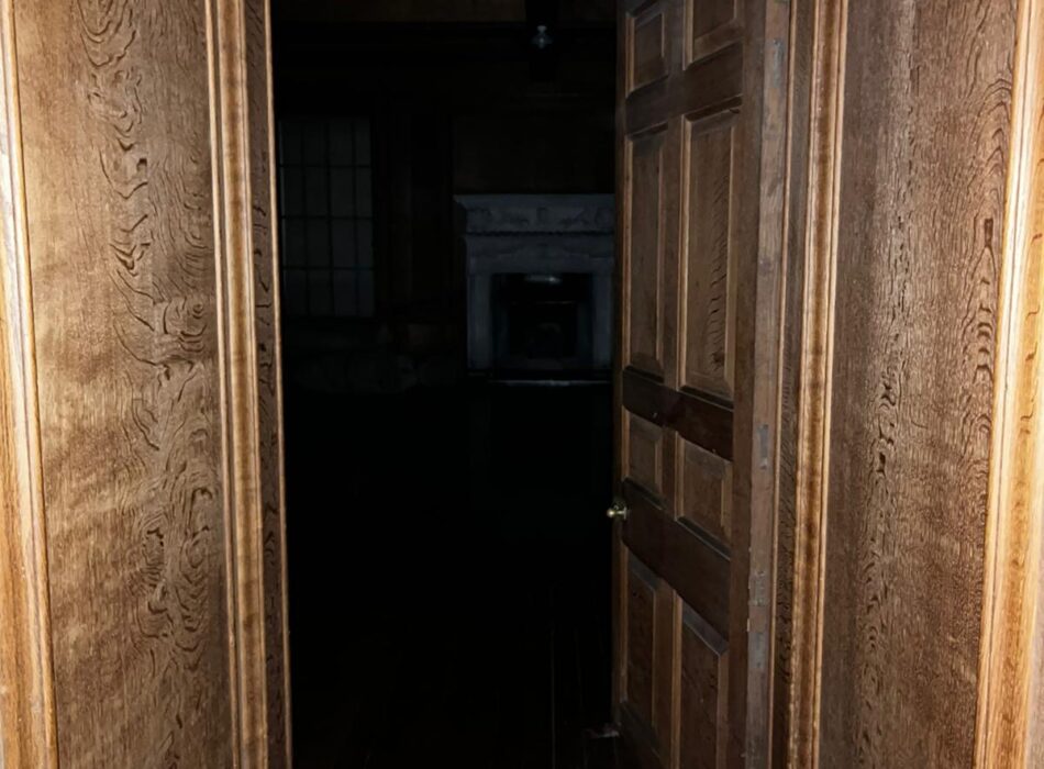 A perspective shot looking through a narrow corridor lined with rich, warm-toned wood paneling and matching dark wood floors. A multi-paneled wooden door stands half-open on the right, leading into a pitch-black room. In the dim background of the unlit room, the faint silhouette of a white decorative fireplace mantel and a gridded window can just be seen.