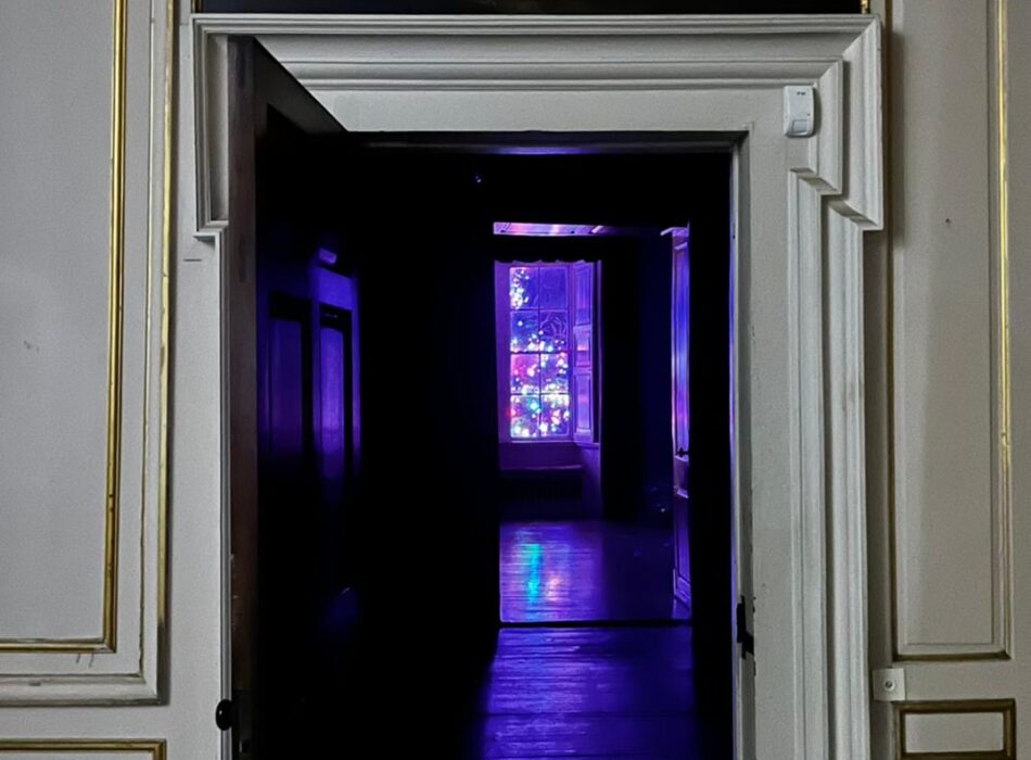 An open, white-paneled doorway leads down a dark corridor toward a distant window illuminated with vibrant purple, pink, and blue lights.