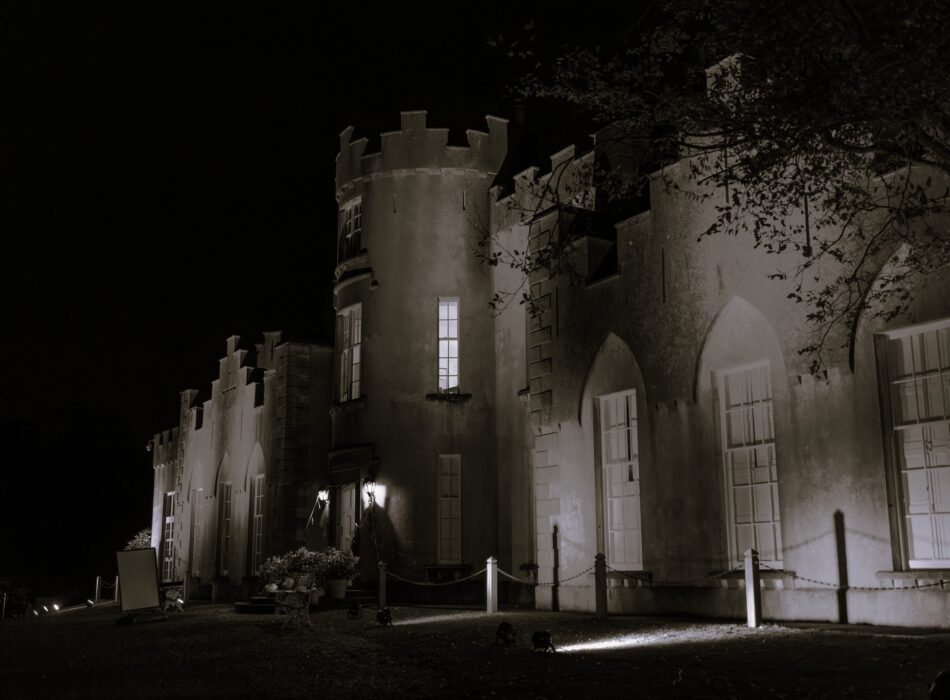 A black-and-white, low-angle shot capturing the front exterior of Ardgillan Castle at night. The structure features prominent crenellated battlements, a circular stone tower with brightly lit windows, and large pointed gothic-style windows on the right. Spotlights placed on the grass patch below cast upward beams of light, illuminating parts of the dark stonework and a nearby tree branch, creating deep shadows.