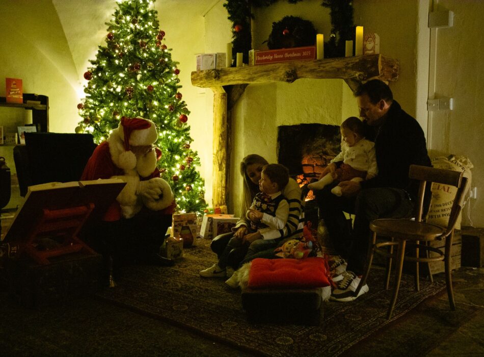A warm, dimly lit festive scene showing a family visiting Santa Claus. On the left, Santa sits beside an illuminated Christmas tree and an open book on a wooden stand. In the center, a mother comforts a young boy on the floor, while a father sits on a wooden chair to the right holding a toddler.