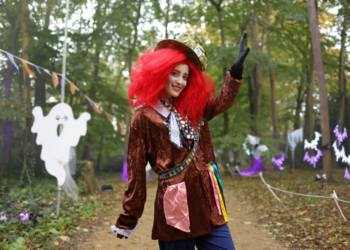 A woman in costume poses before a ghost decoration in the woods. She is wearing a red wig, a red/orange hat and jacket with colourful material coming out of the pockets, blue trousers, a white neck tie and black gloves. One of her arms is in the air while the other is by her side.