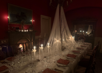 A dining room lit only by candle. It has maroon walls, a chandelier, a fireplace with a fire lighting and a dark dresser. A white table is set in the middle of the room with netting coming down from the chandelier. It has candles, glasses, place settings and dinning menus. There is not yet anyone sitting at the table.