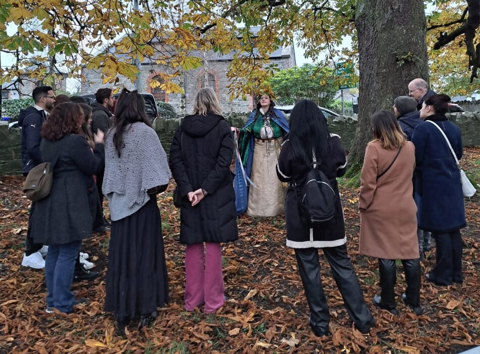 A group of about ten people stands in a circle on a lawn covered in fallen leaves during a Tlachtga Abhaile walking tour in Athboy. They are gathered near a large tree trunk, listening to a female guide dressed in a traditional Celtic costume complete with a leaf crown. A historic stone building and a stone perimeter wall are visible in the background.