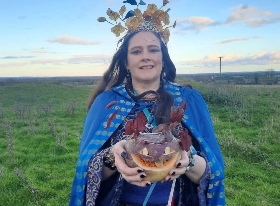 A low-angle shot of a woman portraying Tlachtga, the Celtic Goddess of Halloween. She wears a long blue cape with a patterned trim, a teal gown, and a large headpiece constructed from yellow autumn leaves and berries. She holds a rustic, curved wooden staff and stands outdoors next to a tree trunk against a bright blue sky.