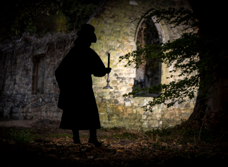 A figure wearing a top hat and black overcoat is seen carrying a candle in front of a faintly lit ruins of a church. On the right-hand side of the picture, a tree can be seen and there are brown leaves in the foreground.