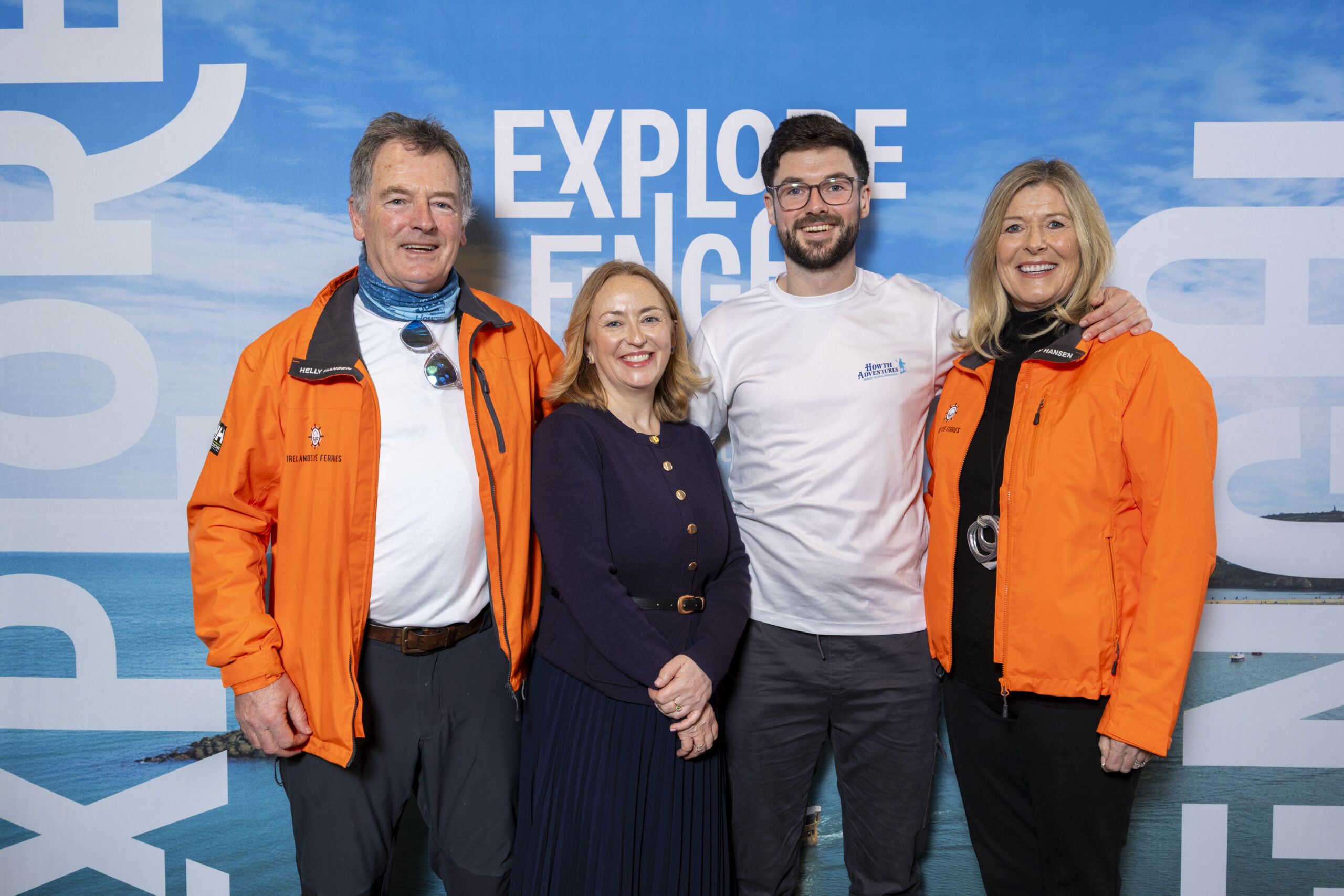 Four smiling people stand in front of an Explore Fingal banner. The man on the left wears an orange jacket, a blue buff, white tshirt and black trousers. Sunglasses hang from his tshirt. The woman beside him wears a professional navy jacket and skirt. The on her right wears glasses, a white tshirt which says 'Howth Adventures' and black trousers. The woman beside him wears an orange jacket, black top and black trousers. They are all smiling at the camera.