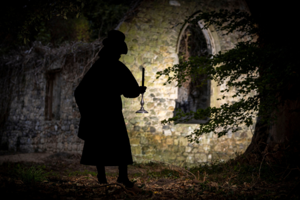 A figure wearing a top hat and black overcoat is seen carrying a candle in front of a faintly lit ruins of a church. On the right-hand side of the picture, a tree can be seen and there are brown leaves in the foreground.