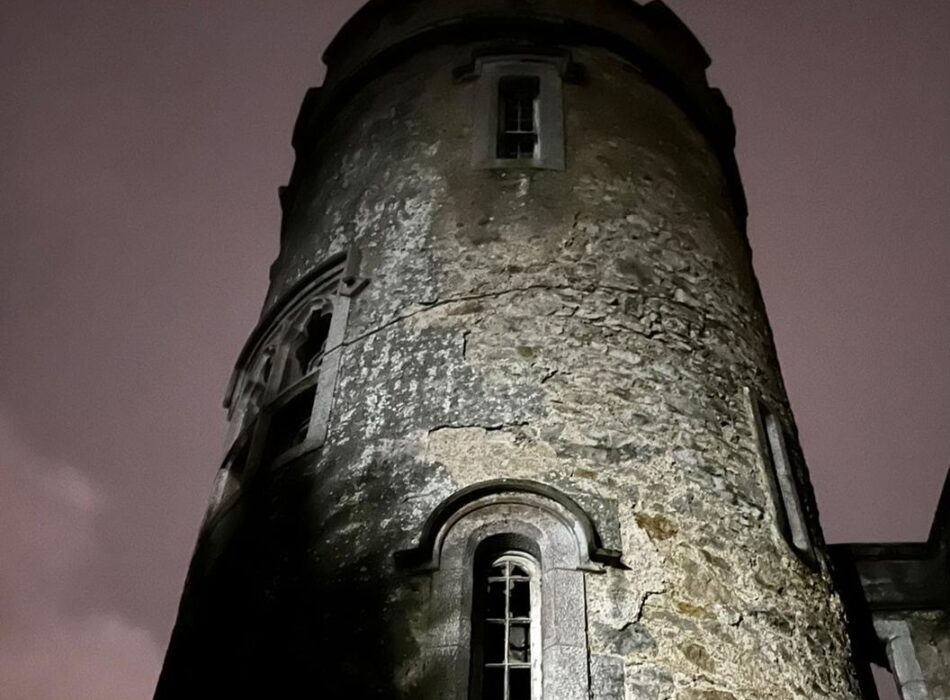 A dramatic low-angle perspective looking up at the circular stone Mermaid’s Tower at Howth Castle at night. The weathered, rustic stone masonry shows cracks and patches of green moss, highlighted by a strong uplight from the bottom left. The tower features a tall, narrow arched window with white frames in the center, a gothic arched window to the upper left, and a smaller rectangular window near the top battlement. The background is a moody, dark overcast sky with a faint pinkish tint.