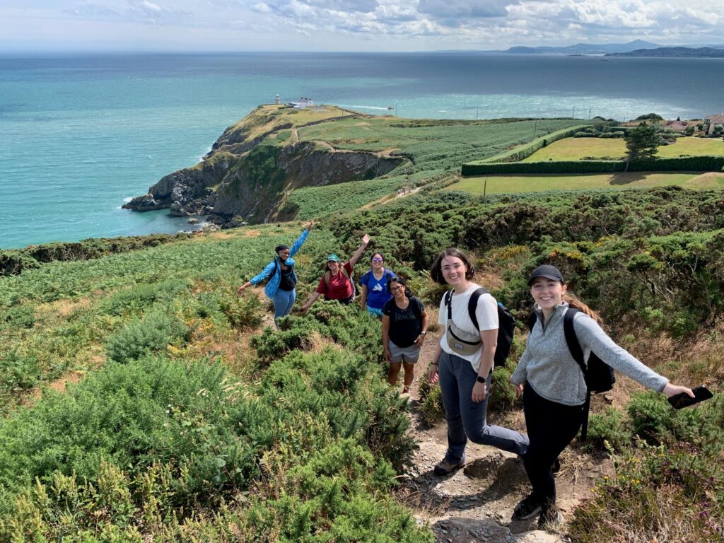 A group of hikers standing on a hilltop along the Howth coastal trail, with a guide and a dog in the foreground and panoramic views of the harbour and sea behind them.