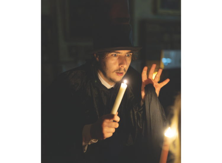 A man wearing a tall hat and dressed in a black cloak and shirt speaks while holding up a candle in a dark room