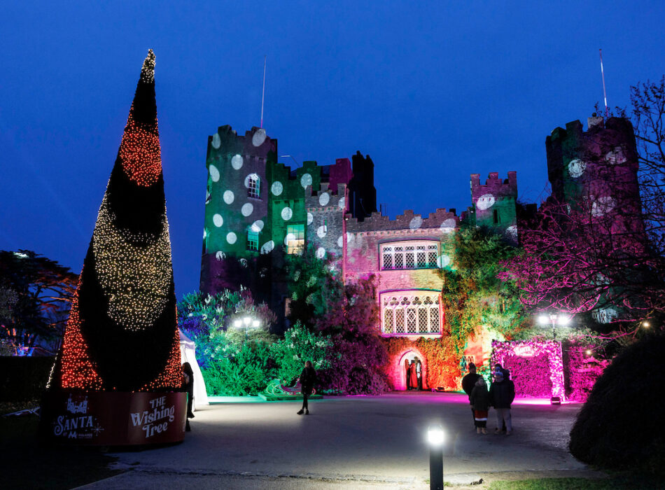 A large stone castle illuminated at night with festive holiday light displays, including green polka-dot and bright pink projections on its walls and towers. In the foreground, a tall, cone-shaped Christmas tree covered in dense fairy lights stands on a base labeled "The Wishing Tree" and "Santa." Several visitors walk around the courtyard under a deep blue twilight sky.