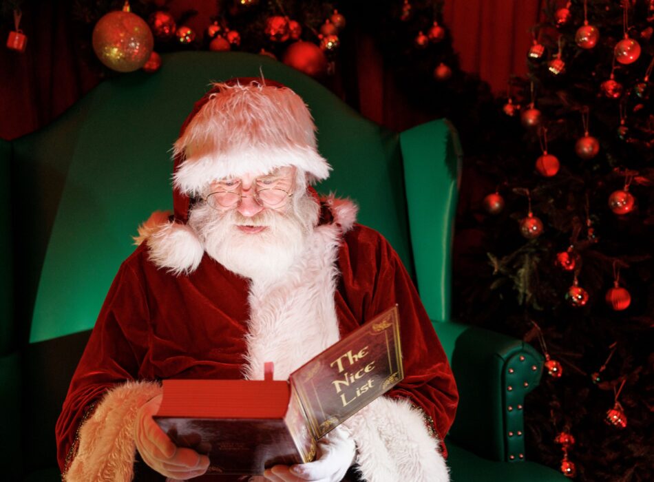 Santa Claus sitting in a large green armchair, wearing his classic red velvet suit and glasses while holding open a book labeled "The Nice List" on the cover. A warm glow shines from the pages onto his face and white beard. The background features deep red tones and a Christmas tree adorned with round red ornaments and warm festive lighting.