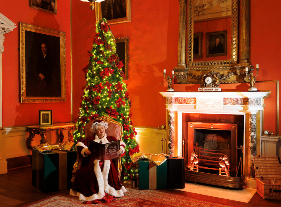 Mrs. Claus sitting in a grand armchair inside an elegant room with deep orange-red walls, reading a book titled "Santa Stories." Next to her stands a tall, heavily decorated Christmas tree lit with warm fairy lights and red ribbons, surrounded by large wrapped gifts. To the right is a classical marble fireplace with an overhead golden mirror, antique paintings on the wall, and architectural pillars.