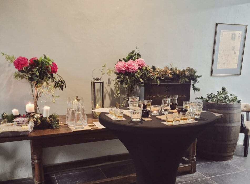 A high-top tasting table arranged with whiskey flight glasses next to a wooden side table decorated with pink flowers and candles.