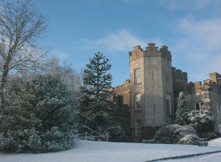Ardgillan castle is on the right-hand side of the image while the left-hand side of the image shows lots of tree, both bare deciduous trees and evergreen trees. There is a bright blue sky with a few soft clouds and the grounds of the castle are covered in snow.