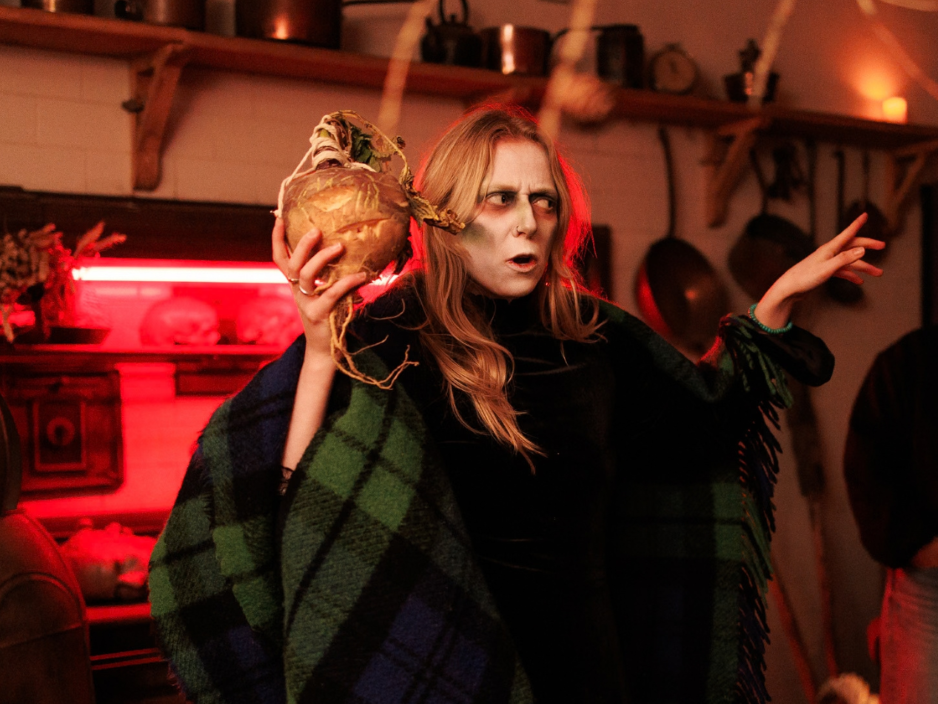 An actor wearing a green tartan cloak with facepaint tells a story in a kitchen while holding a turnip.