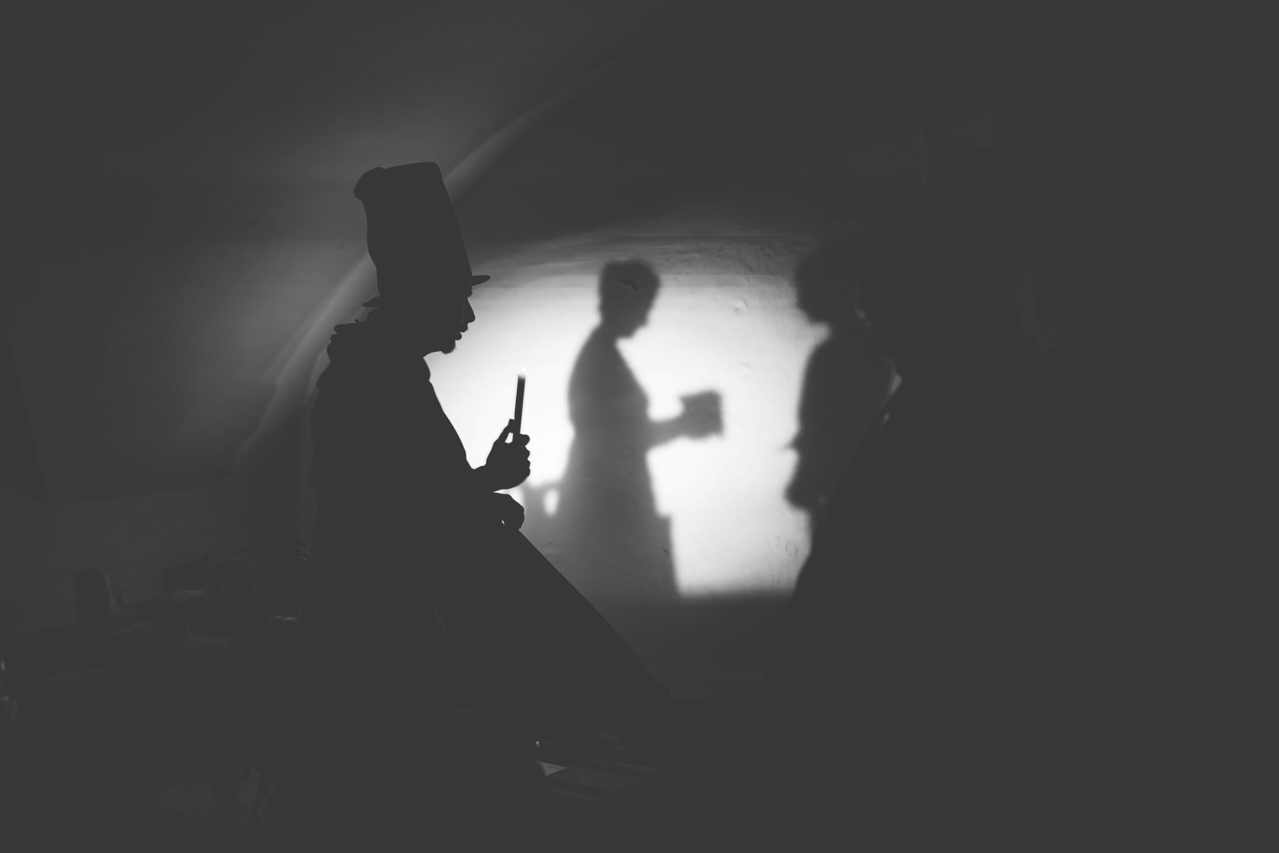 A photo of a dark room with one lamp shining light on the far wall. There is the silhouette of a man in a top hat holding a candle, and the shadow on the far wall is a woman looking down at a small box in her arms. A second shadow on the wall can only be partially seen and it looks at the box as well.