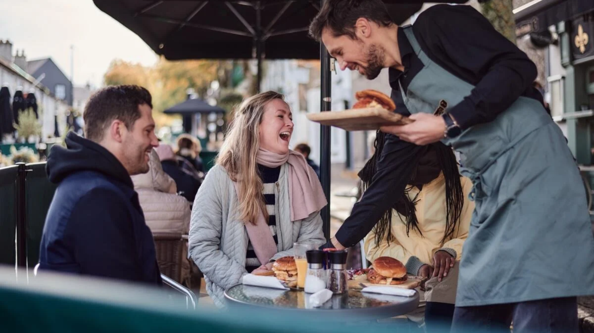 Three friends sit at an outdoor table being served food by a waiter. The outdoor table is on a town street, covered by an umbrella and more tables are visible in the background. The three friends in the picture are laughing and wearing coasts and scarves. One of the friends is blocked by the waiter who leaves burgers on the table.