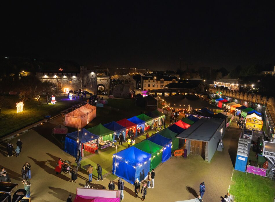 A high-angle night photograph of an outdoor Christmas market inside a castle courtyard. Rows of market stalls with illuminated blue, green, and red canopy tents are lined up on the grounds. Visitors walk along the pathways between stalls, and festive light displays, including a glowing Santa figure, decorate the grassy areas near the historic stone walls.
