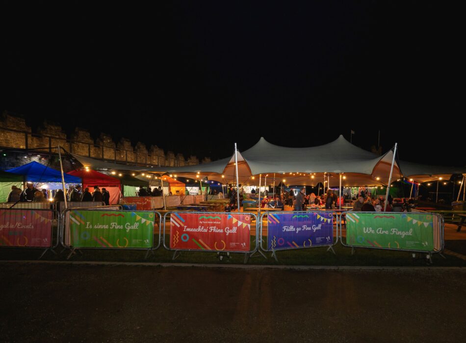 A night photograph of an outdoor event space at a Fingal market. A large white stretch tent covers a seating area with picnic tables. In the foreground, metal crowd barriers are covered with colorful promotional banners featuring Irish and English text like "We Are Fingal" and "Is sinne Fine Gall." In the background, illuminated vendor stalls line a historic stone wall.